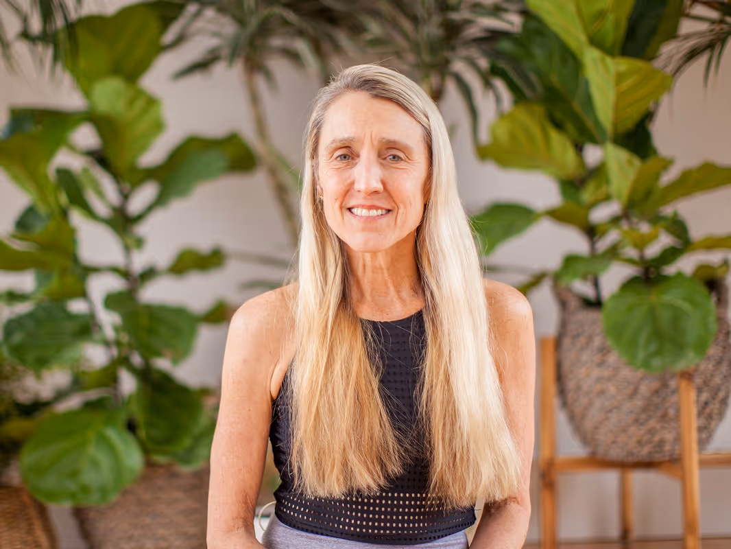 Smiling middle-aged woman with long blonde hair wearing a black sleeveless top, standing indoors with large green plants in the background.