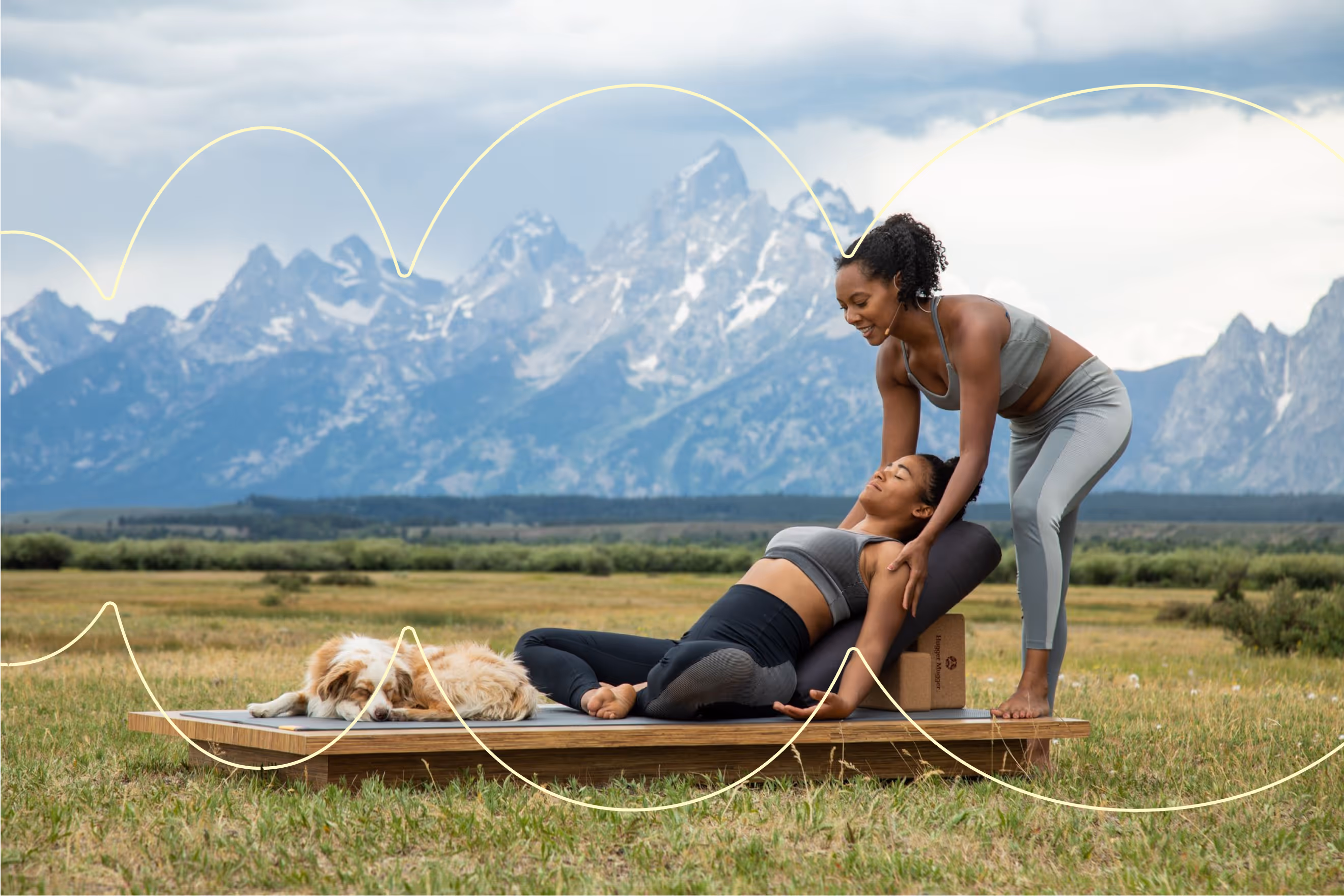 Woman assists another doing yoga on mat outdoors near sleeping dog with mountains in background.
