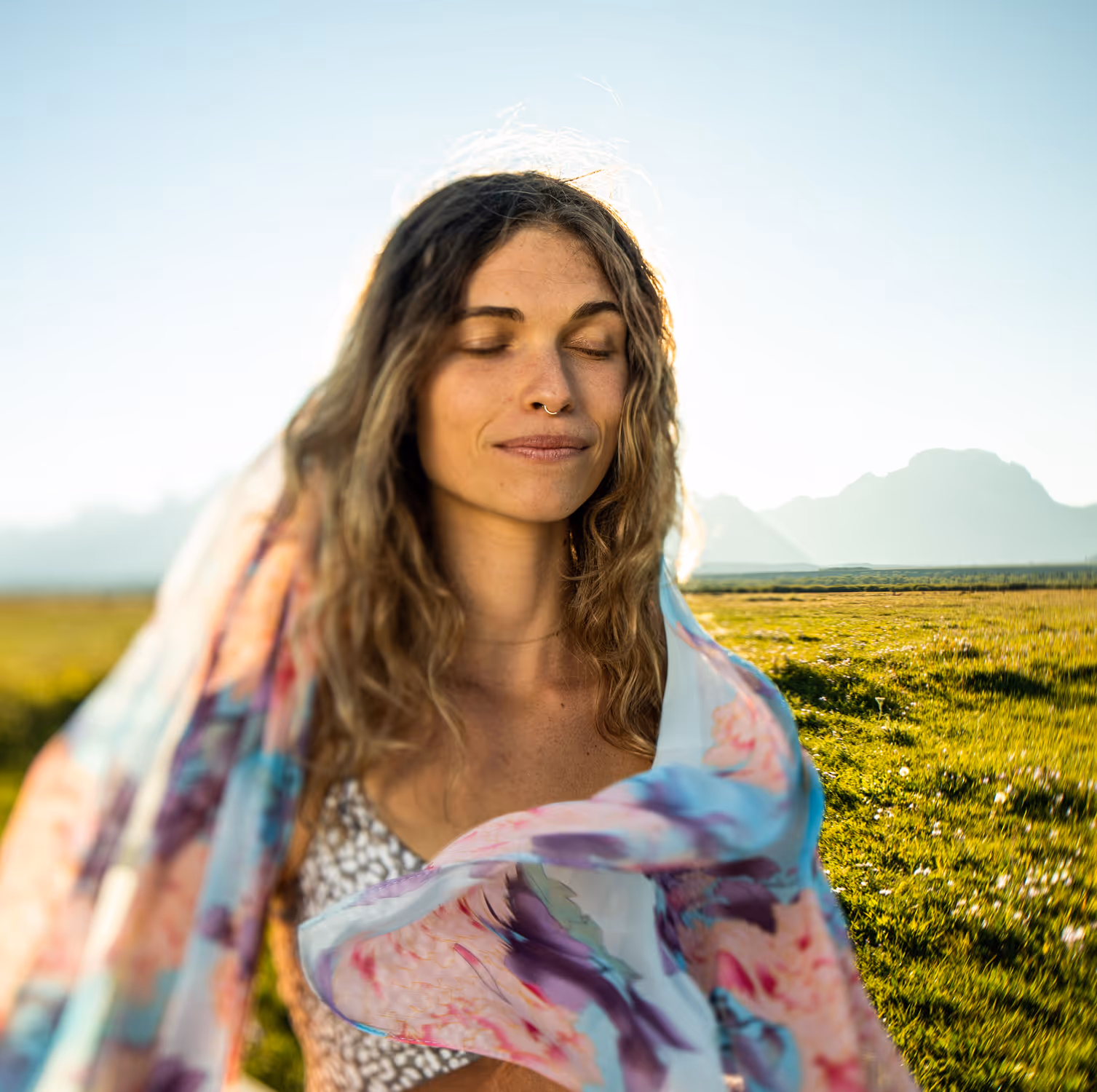 Woman with closed eyes and flowing scarf stands in a sunny grassy field with mountains in background.
