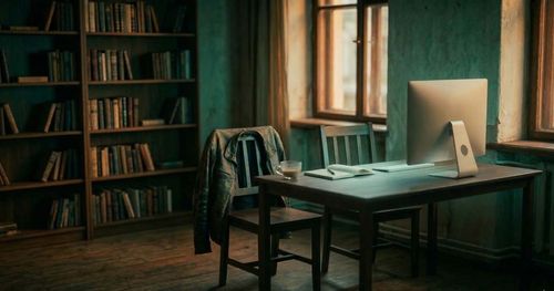Desk in a home office shows one empty chair with a jacket and nearby coffee cup and notebook, and a second empty chair in front of a keyboard and computer monitor