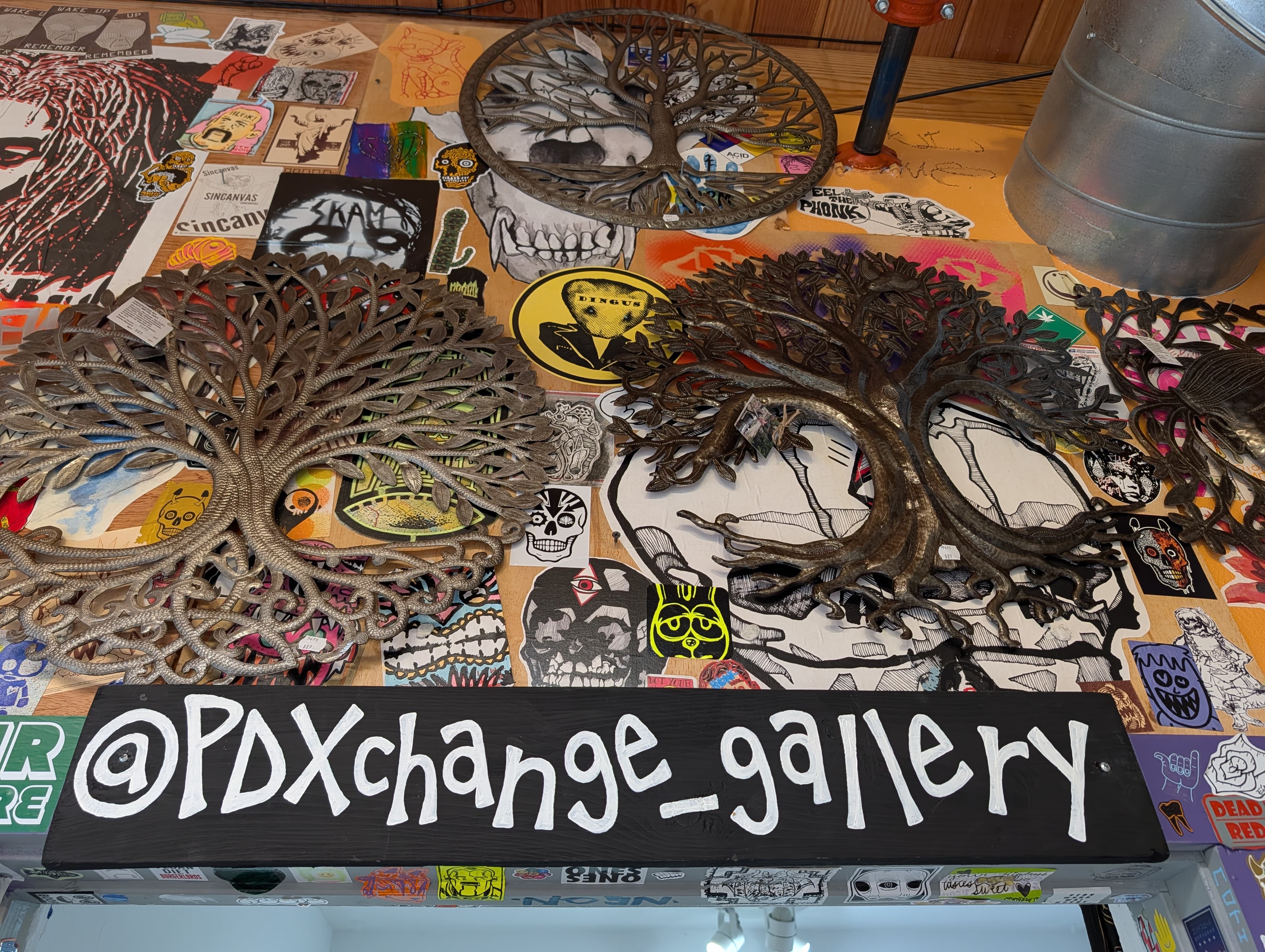 PDXGallery's Sign, insta handle @pdxchanage_gallery over a walll covered in stickers and metal art.