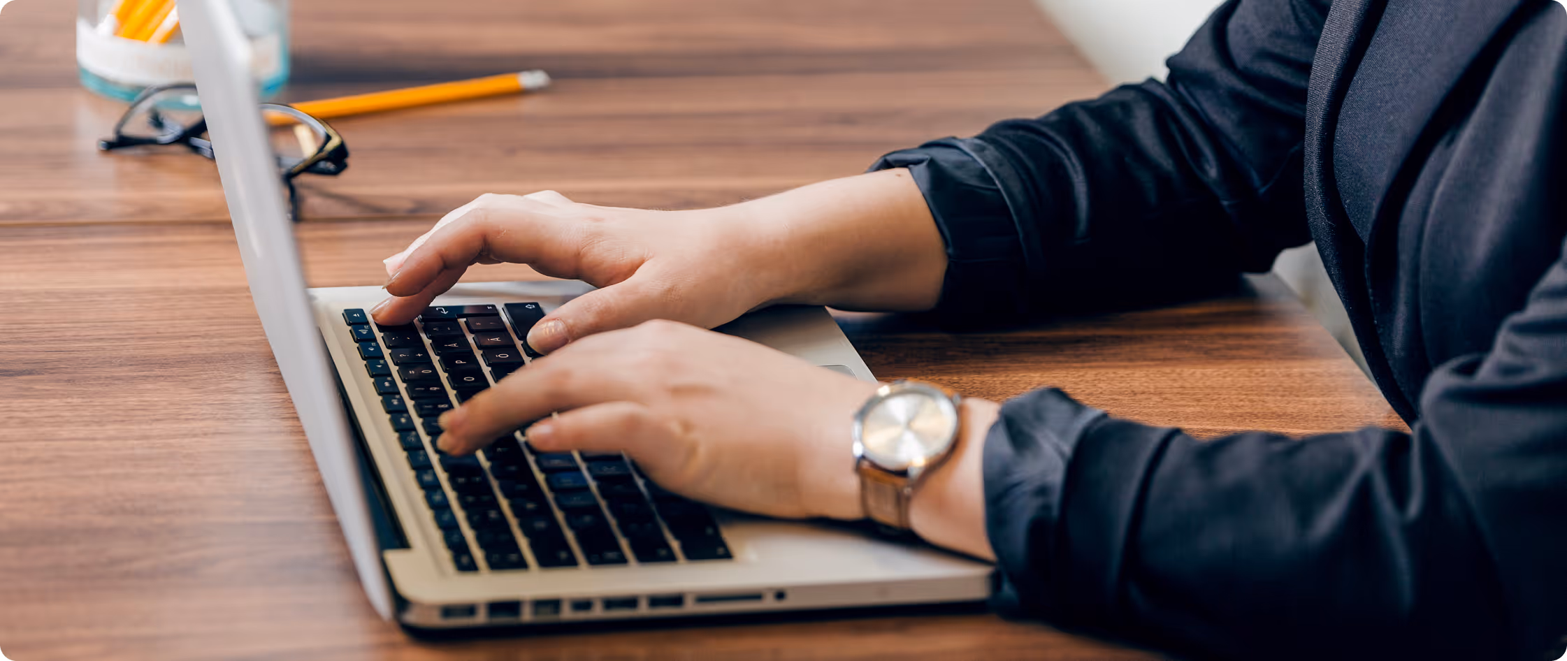 Person typing on a laptop keyboard at a wooden desk with glasses and pencil nearby.