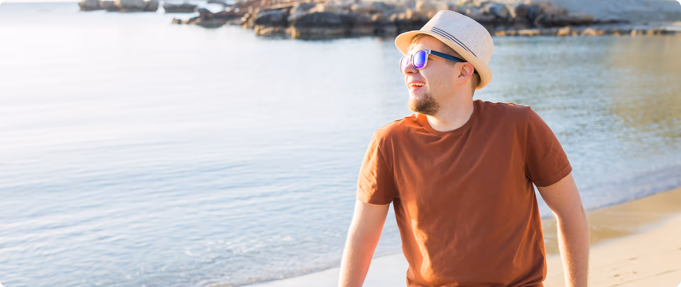 Young man wearing sunglasses and a hat smiling by the water at a sunny beach.