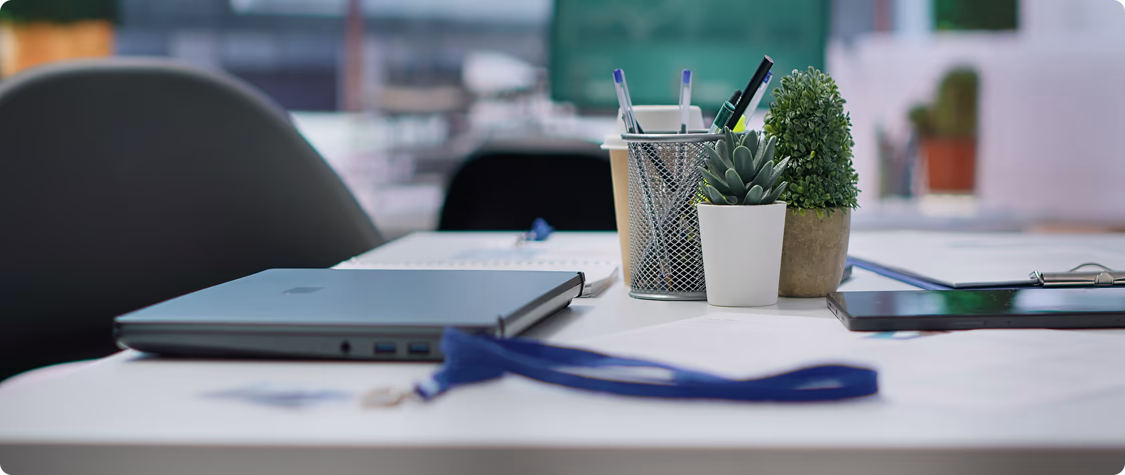 Office desk with a closed laptop, pen holder, two potted plants, papers, and a blurred chair in background.