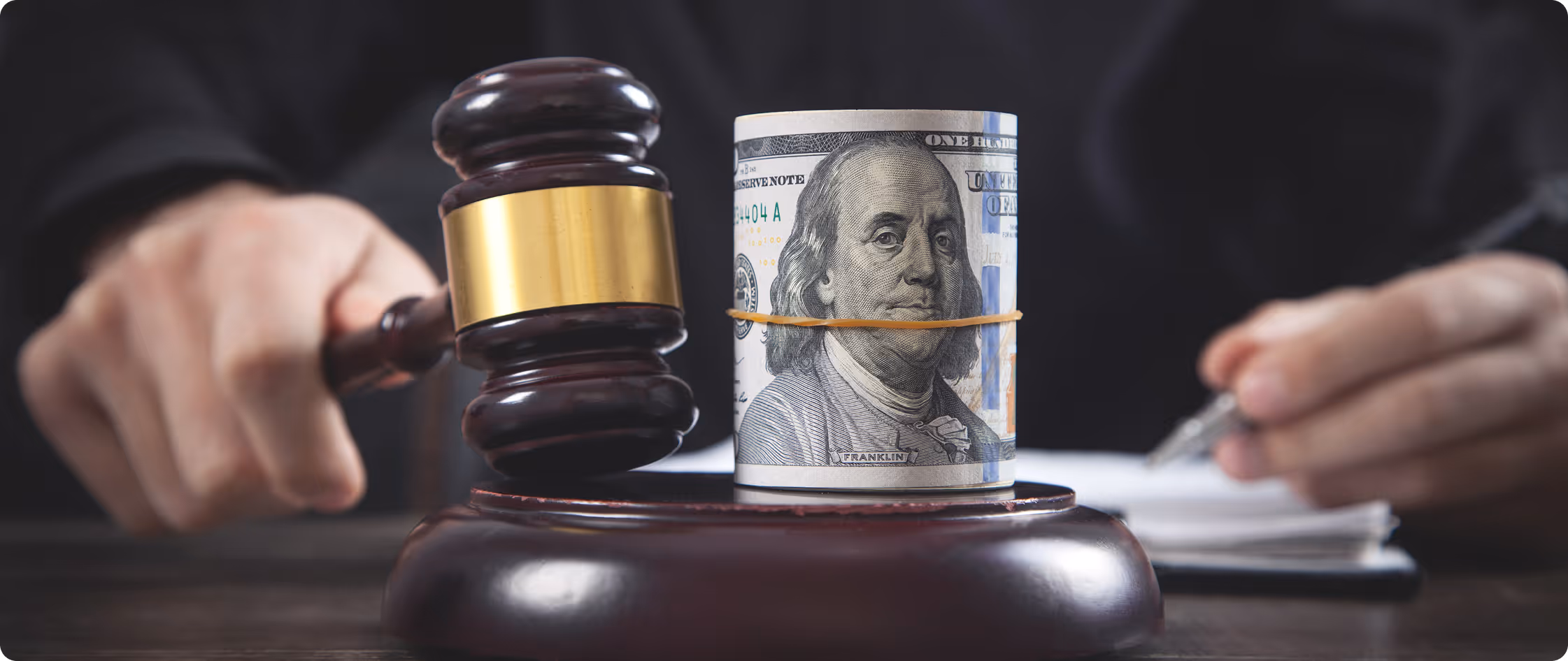 Judge's gavel striking block with rolled 100-dollar bill bound by rubber band on top.