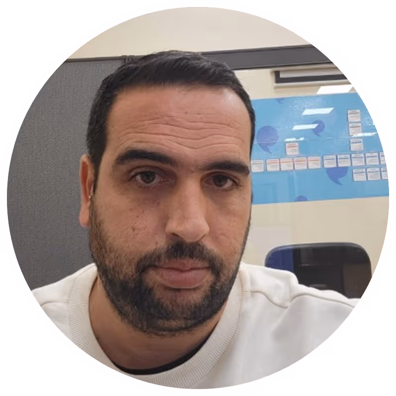 Man with short dark hair and beard wearing a white sweater sitting indoors by office glass wall.