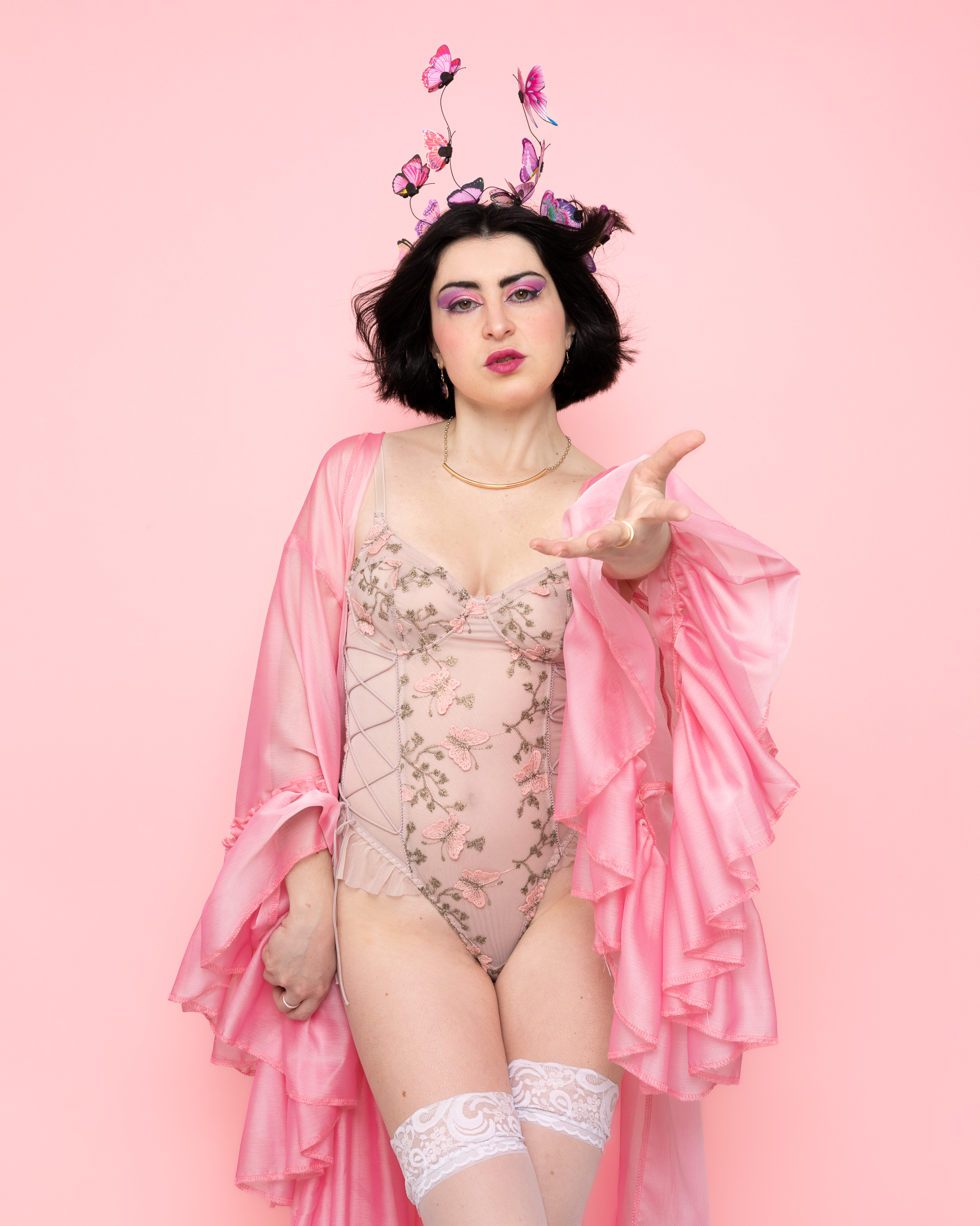 Femme avec maquillage violet, portant une tenue rose fleurie et un kimono rose, avec une couronne de papillons sur fond rose, tendant la main.