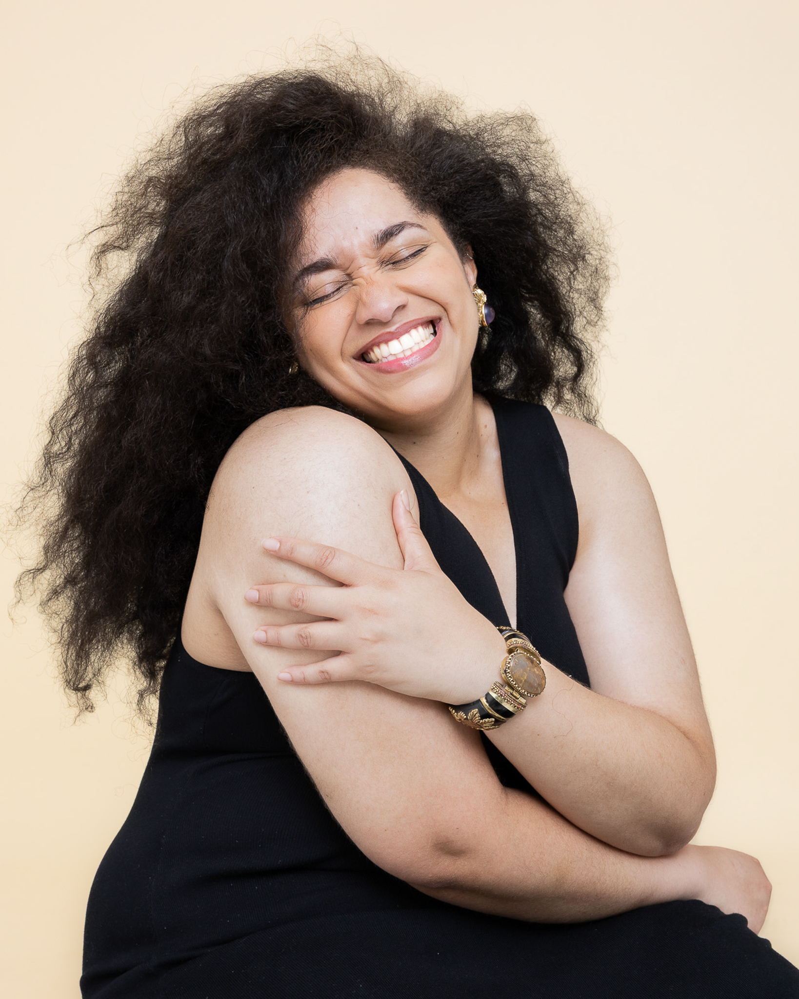 Femme souriante aux cheveux bouclés portant une robe noire, serrant son bras avec joie.
