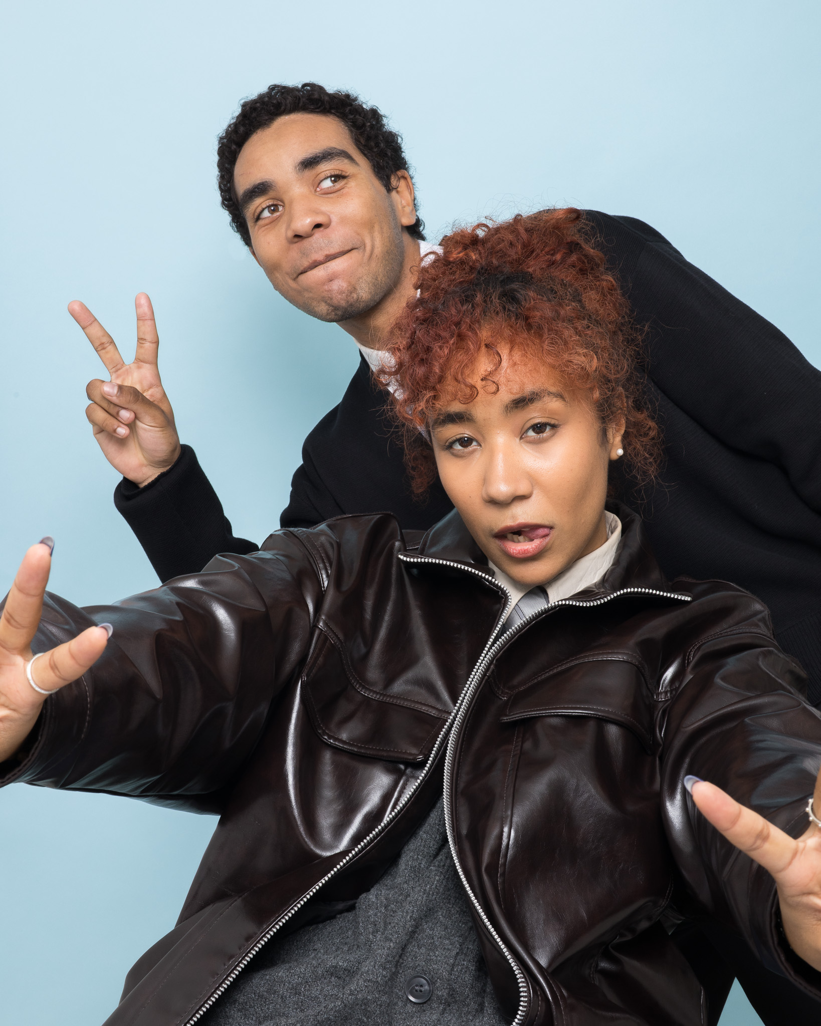 Deux jeunes adultes posant avec des gestes de paix et des expressions faciales joyeuses sur fond bleu clair.