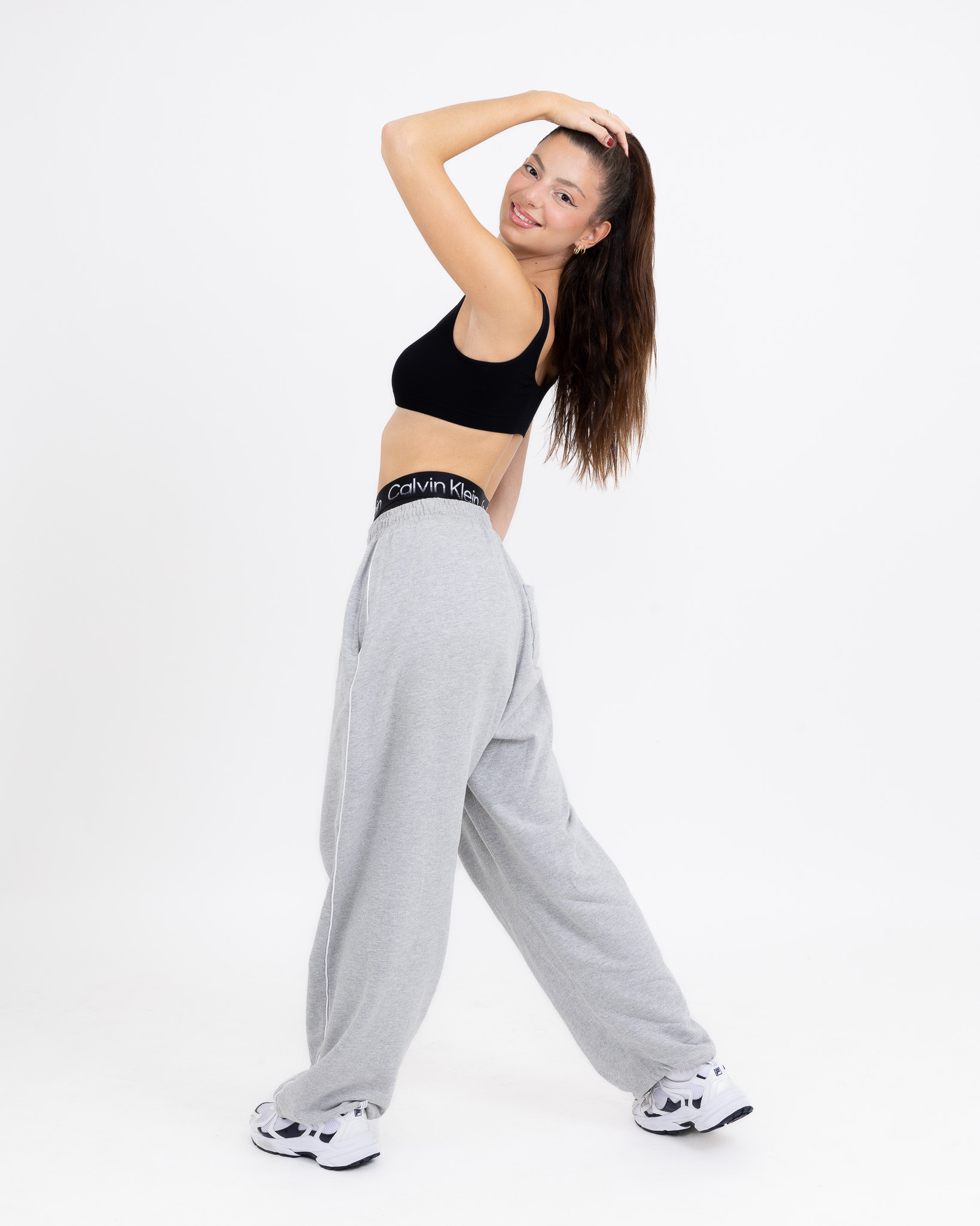 Jeune femme souriante en tenue de sport décontractée, posant avec une main dans les cheveux sur fond blanc.