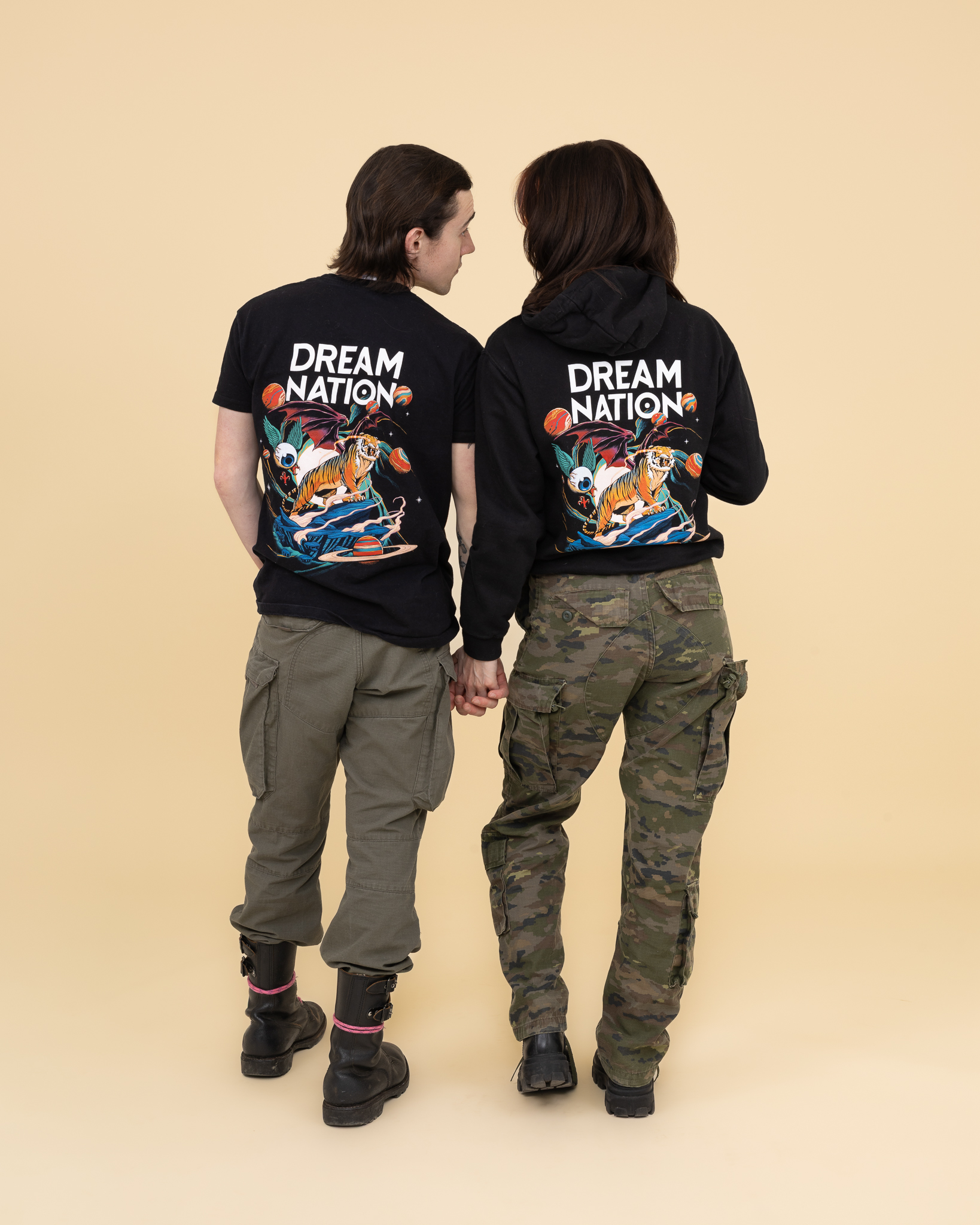 Deux personnes se tenant la main, vues de dos, portant des vêtements noirs avec un motif coloré de tigre sur fond spatial et le texte « DREAM NATION ».