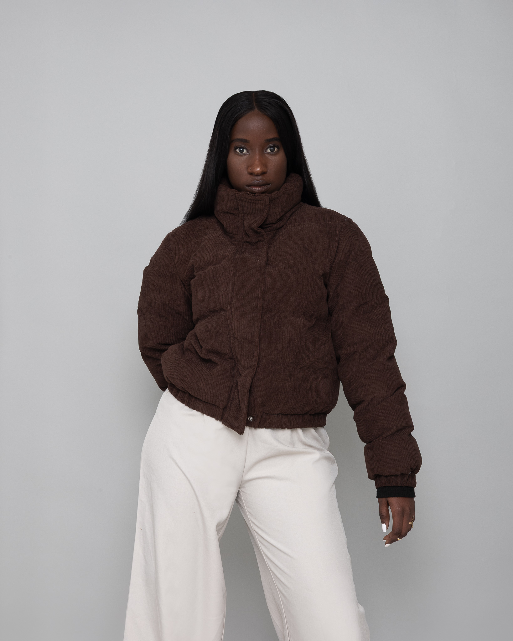 Femme noire portant une veste matelassée marron avec un col montant et un pantalon large blanc, posant devant un fond gris.