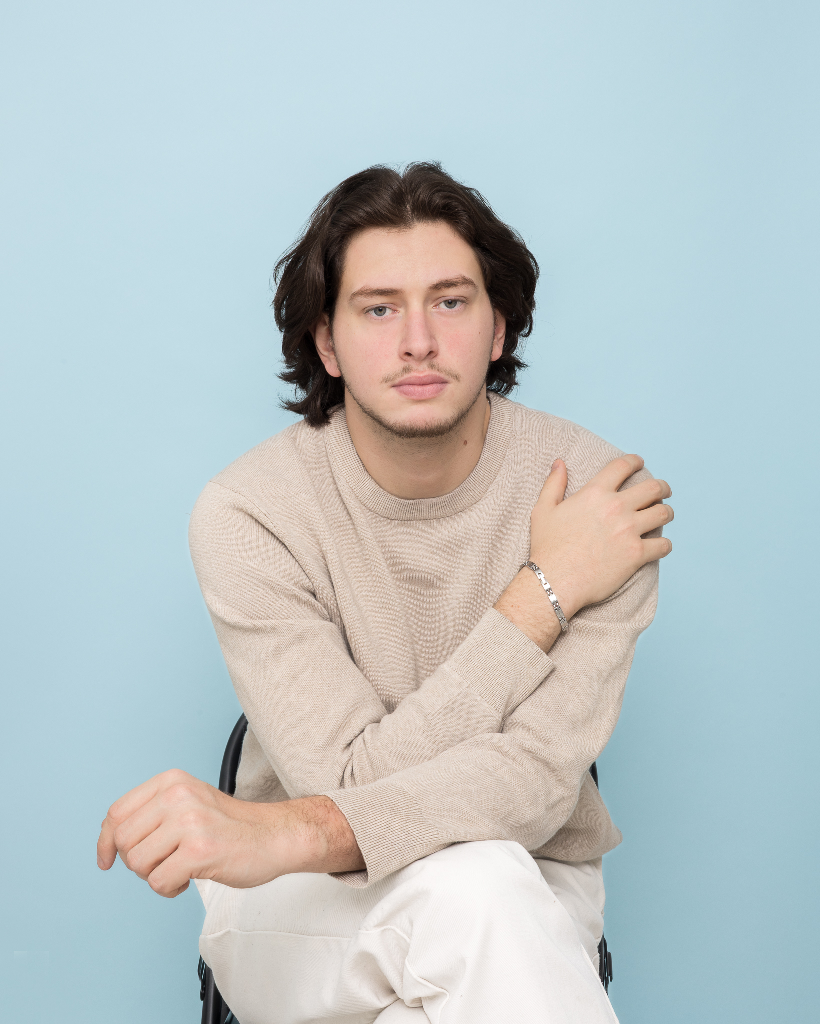 Jeune homme aux cheveux longs portant un pull beige et un bracelet en métal, assis sur une chaise devant un fond bleu clair.
