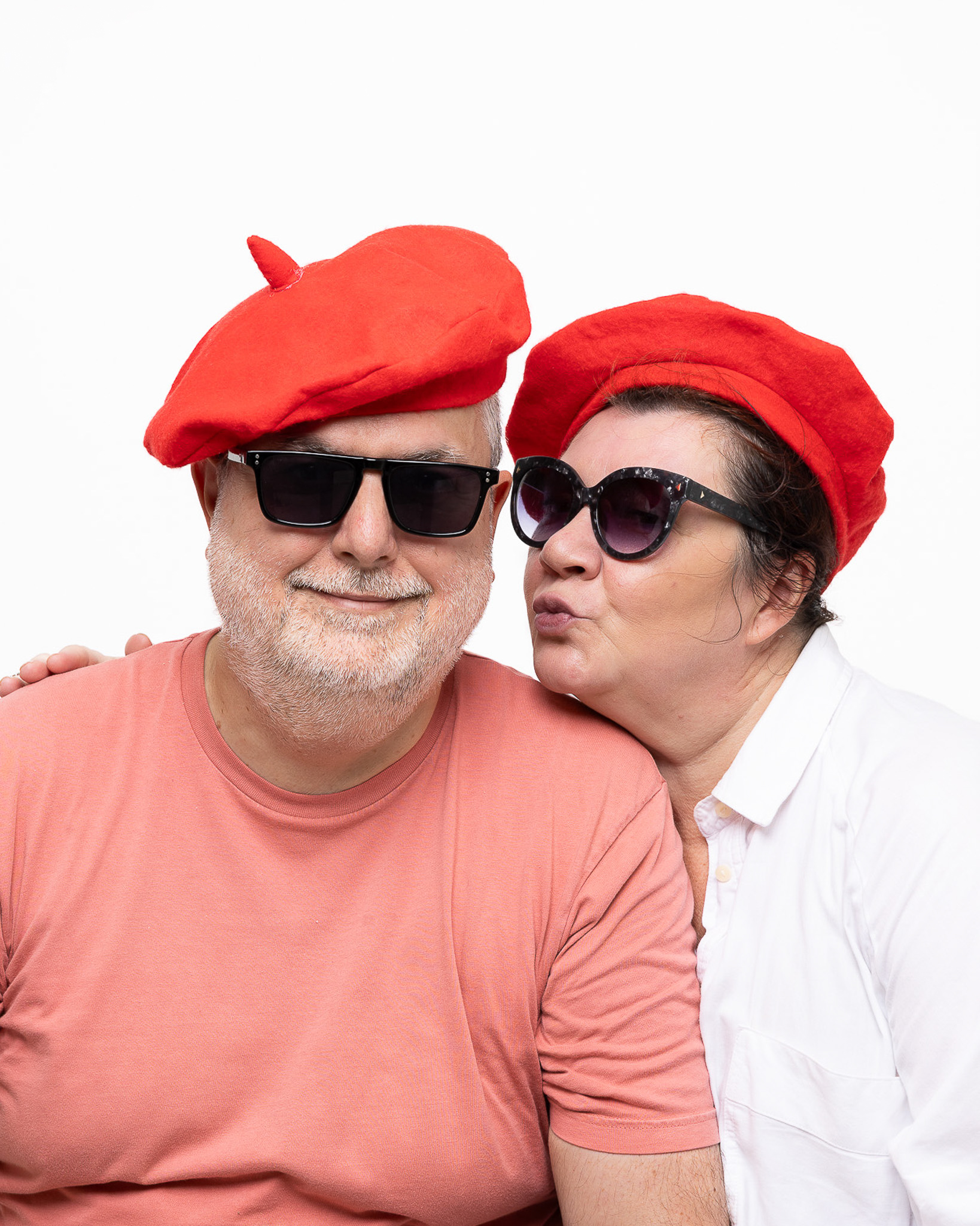 Un homme souriant et une femme lui faisant un bisou sur la joue, portant tous deux des bérets rouges et des lunettes de soleil.