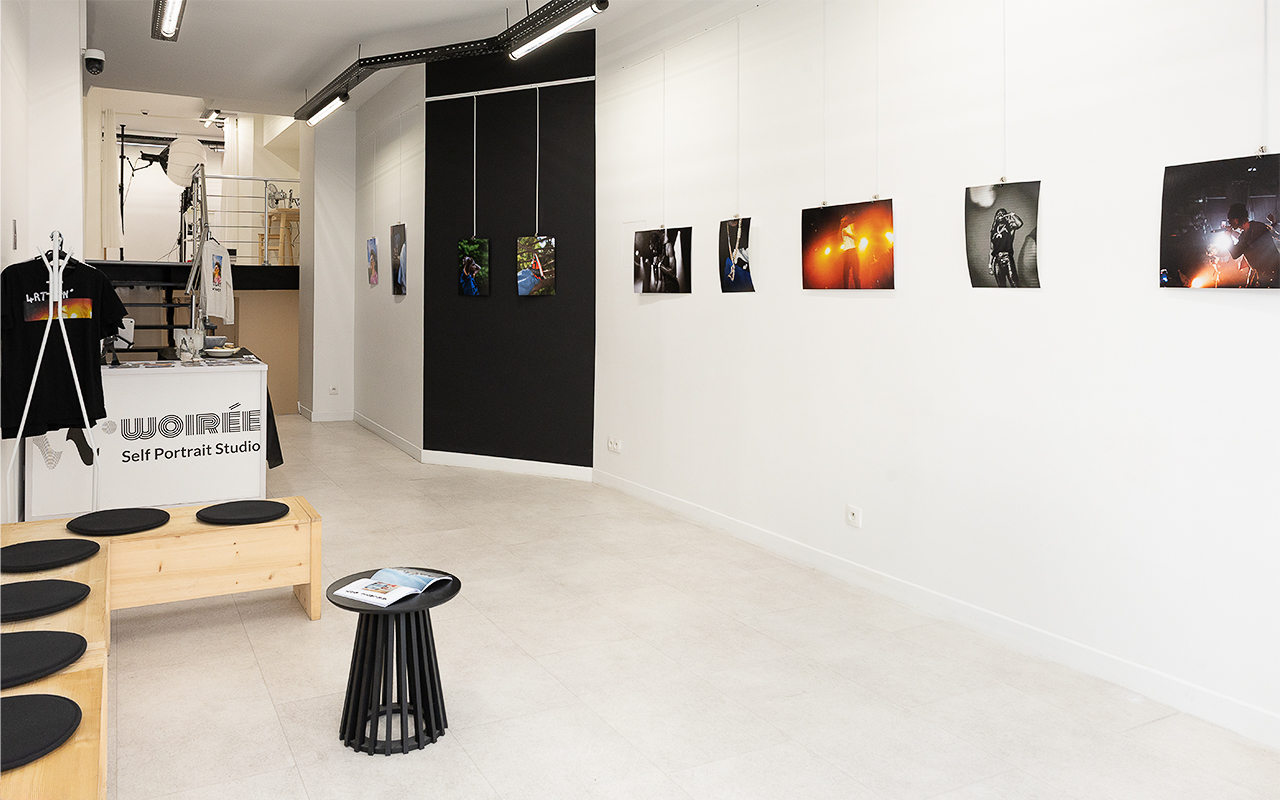 Galerie lumineuse avec photographies accrochées aux murs blancs et bancs en bois avec coussins noirs.