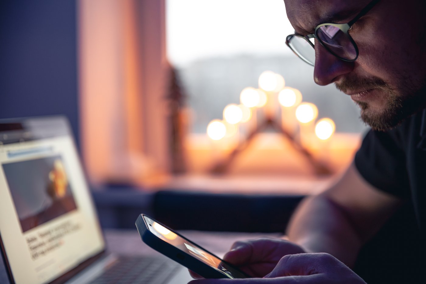 Person mit Smartphone in der Hand vor einem Laptop am Abend