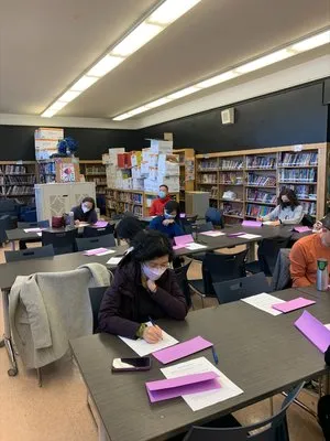 girl imageAdults seated at tables in a library classroom writing on pink cards.