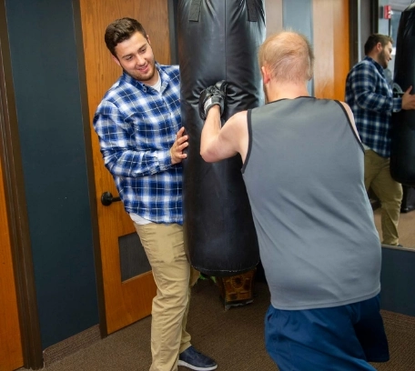 Resident and caregiver at Flatrock engaging in fun boxing exercise on heavy bag in accessible gym amenity, promoting healthy independent lifestyles for individuals with care plans.
