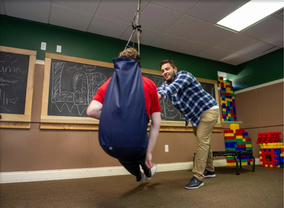 Flatrock sensory room with caregiver assisting resident in suspended therapy swing for relaxation and sensory engagement, supporting autism and well-being.