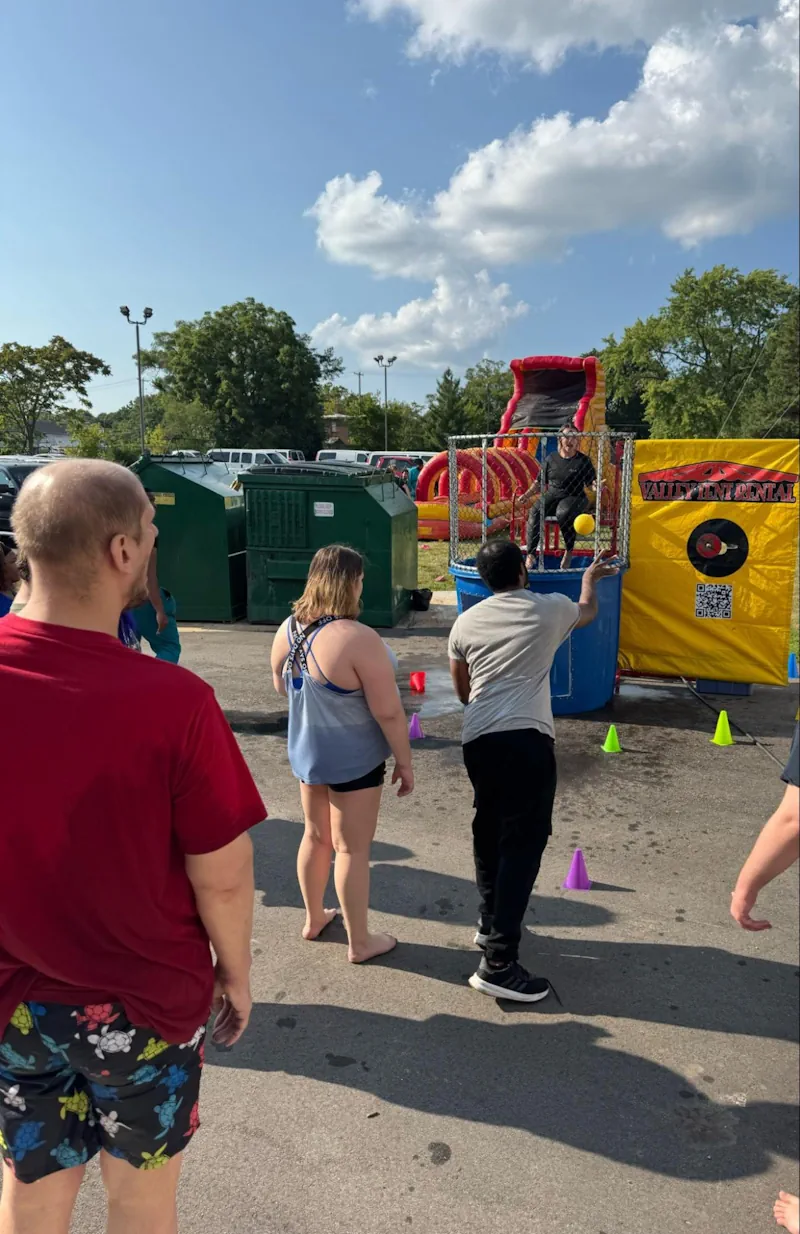 Group of people watching a participant throw a ball at a dunk tank target during a sunny outdoor carnival, with colorful setup and casual attendees enjoying the fun activity.