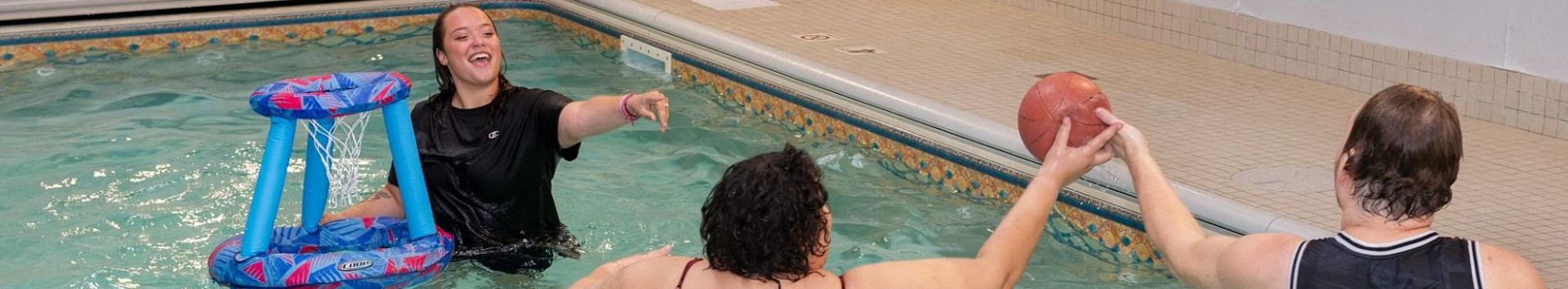 Group playing water basketball with a floating hoop in a spacious indoor swimming pool.