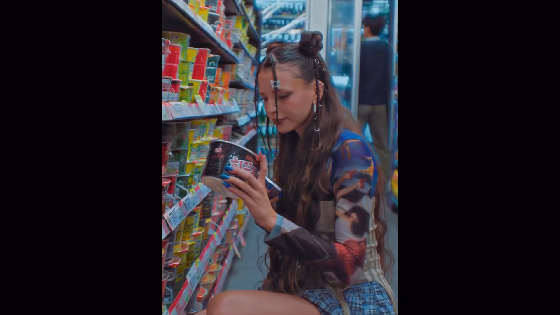 편의점 매대 사이에 앉아 컵라면을 고르고 있는 니나 츄바. Nina Chuba sitting between convenience store shelves, choosing a cup ramen.