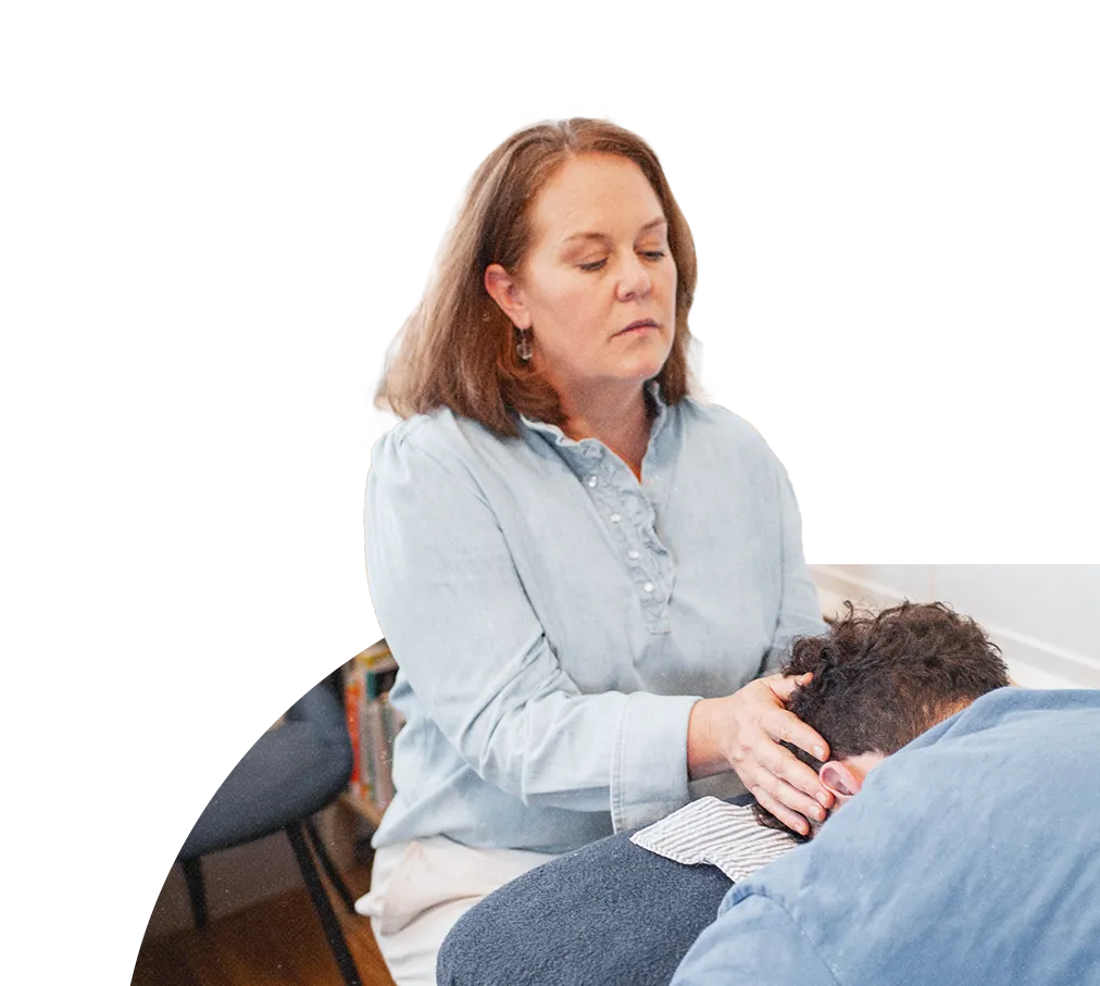 A practitioner in a light blue shirt performing a Spinal Flow treatment, gently placing hands on a client's temple while they lie face down on a cushioned surface.
