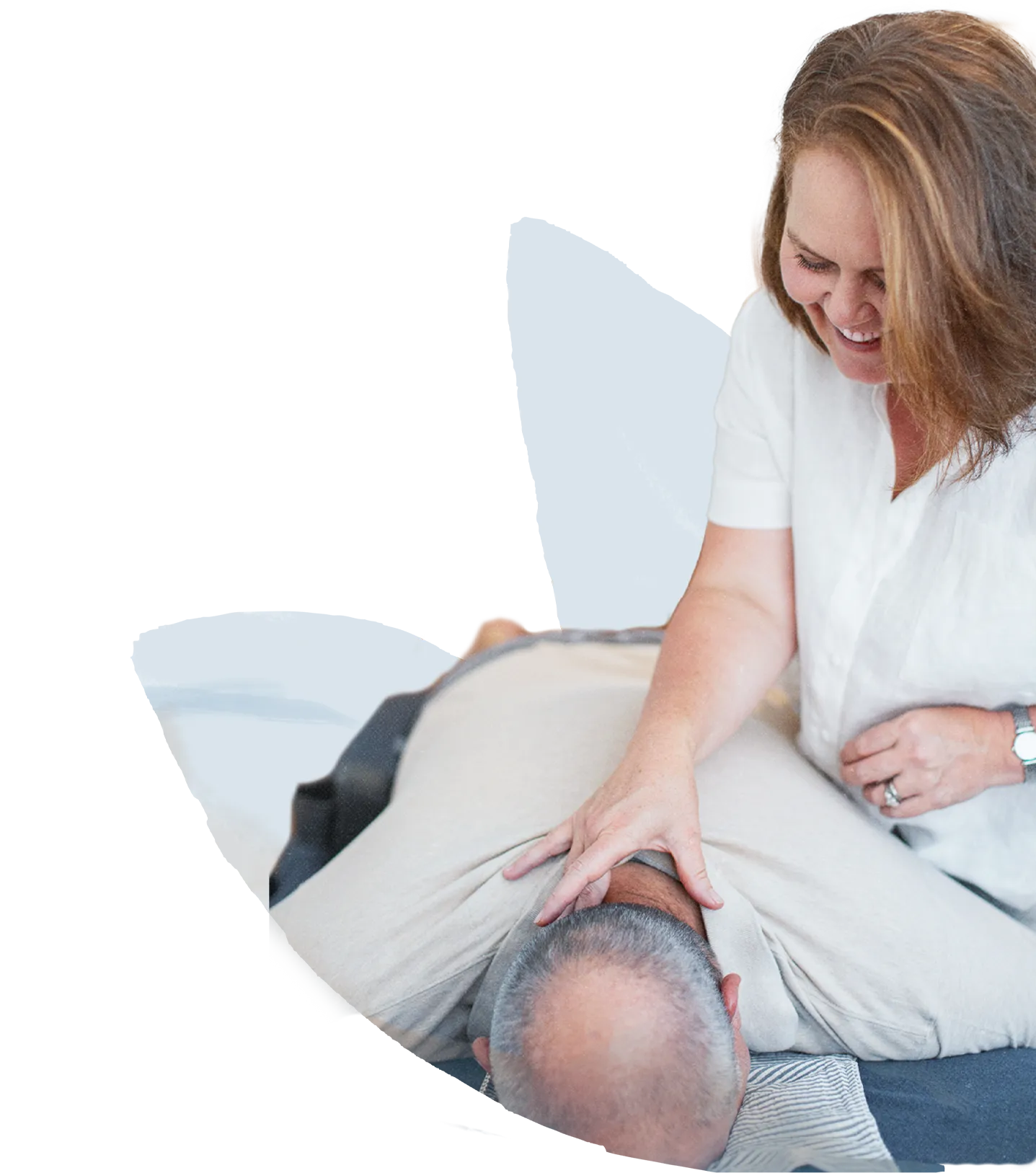 A Spinal Flow practitioner in a white shirt smiling while applying gentle touch to a client's neck and upper back as they lie face down on a mat.