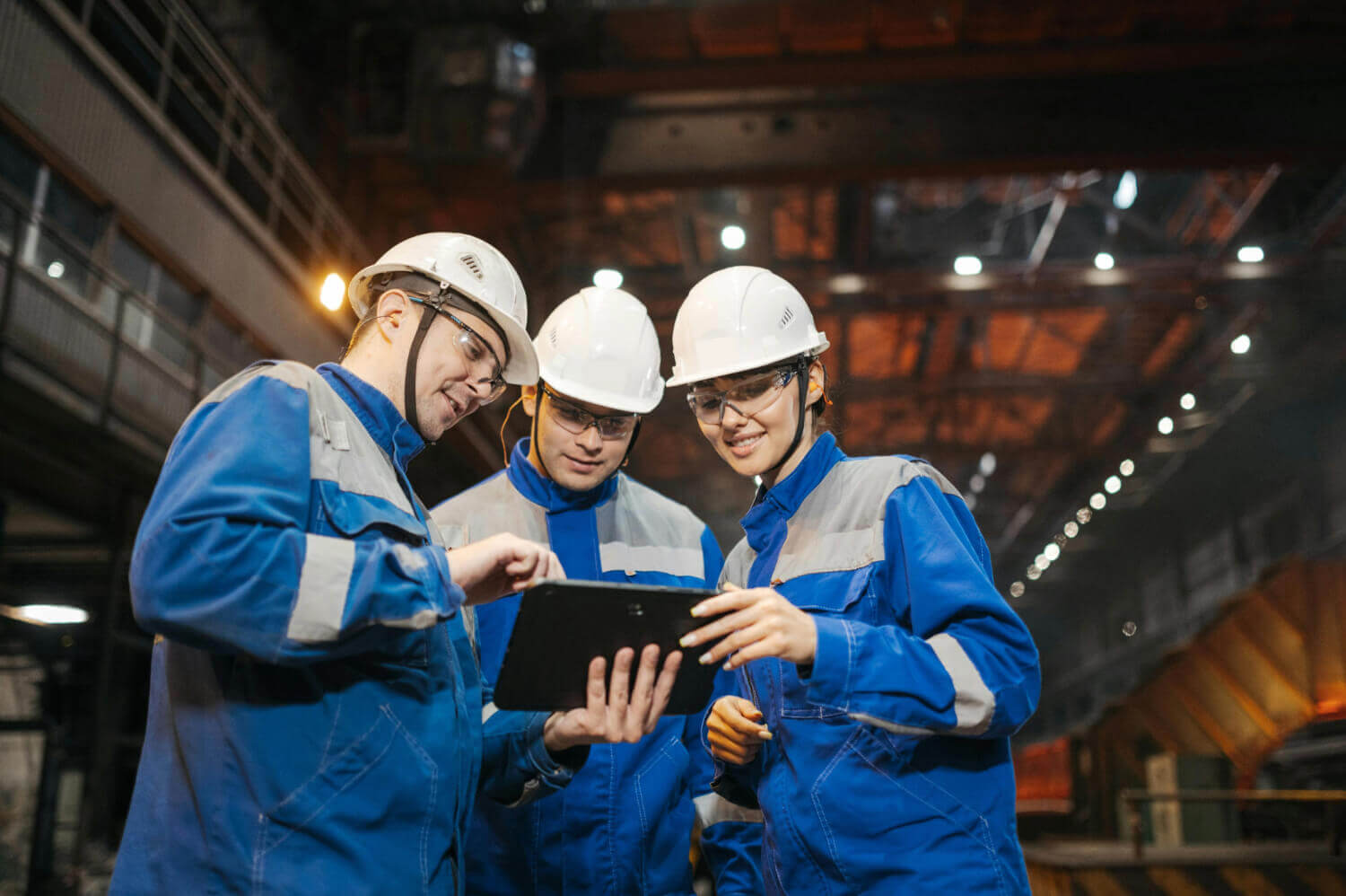 Three construction workers in protective gear and helmets discuss construction machinery financing on a job site using a tablet.