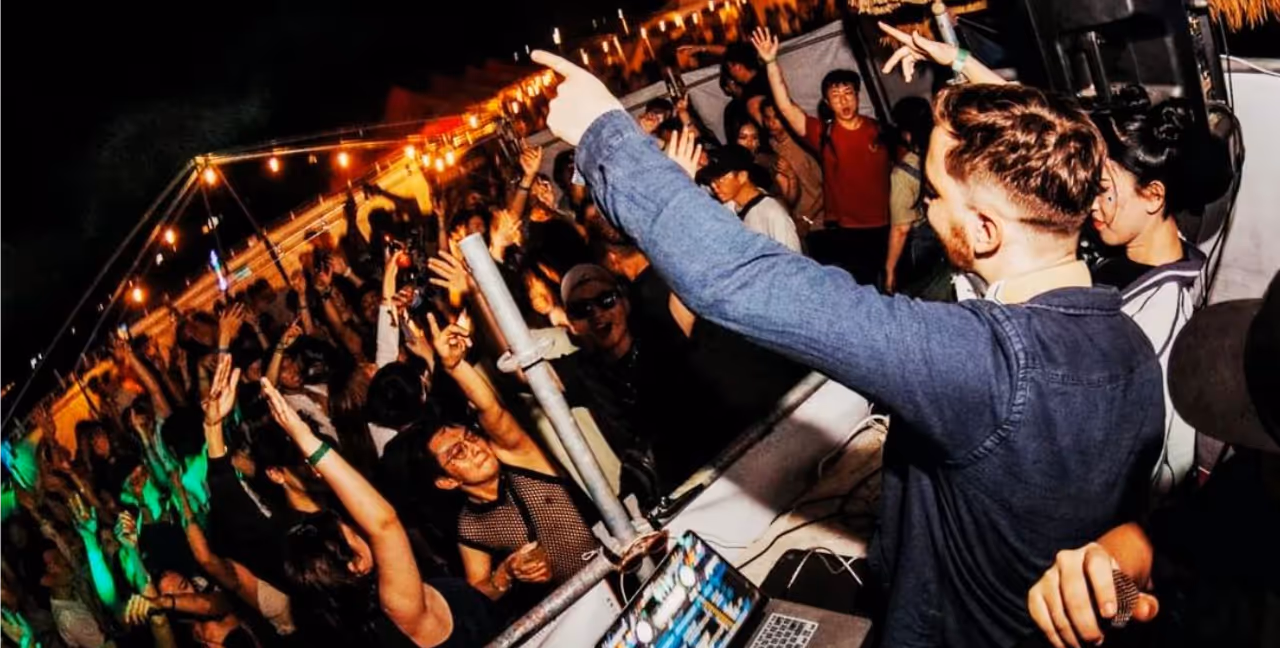 Azido 88 performing in front of a lively crowd at an outdoor event, with people dancing and raising their hands in the air. He is focused on his DJ equipment, with festive string lights and a vibrant atmosphere adding to the energetic scene.