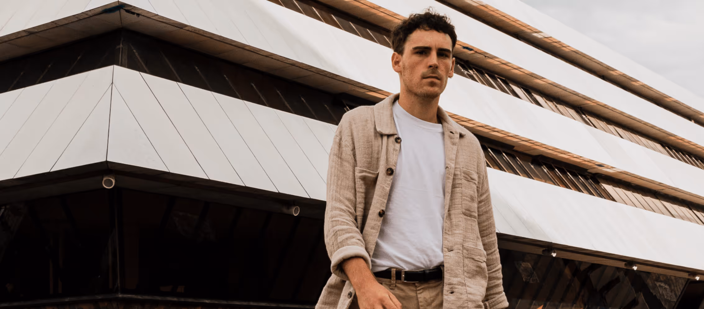 Nhyx standing in front of a modern, angular building, wearing a beige jacket over a white t-shirt. His expression is serious and focused, blending with the architectural lines of the structure behind him.