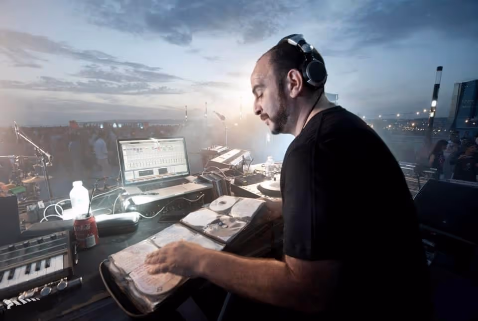 DJ Oil performing at an outdoor event, focused on his equipment with a laptop and various music gear around him. He wears headphones and a black shirt, with a crowd visible in the background under a dusky sky, creating an atmospheric scene.
