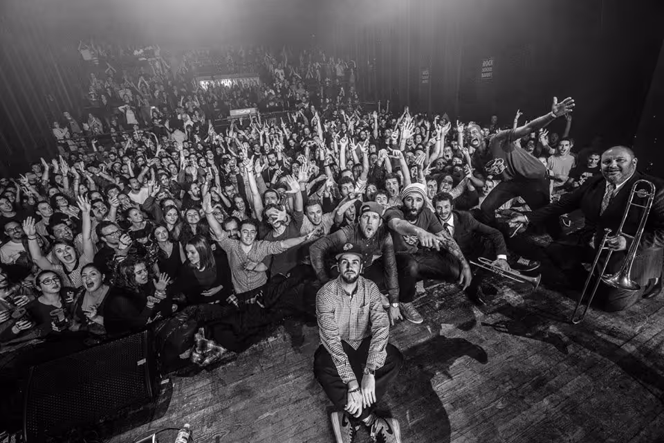 Smokey Joe & The Kid posing with their audience after a performance, capturing an energetic and joyful moment. The band members, along with their fans, are all smiling and cheering, creating a lively and communal atmosphere in this black-and-white photo.