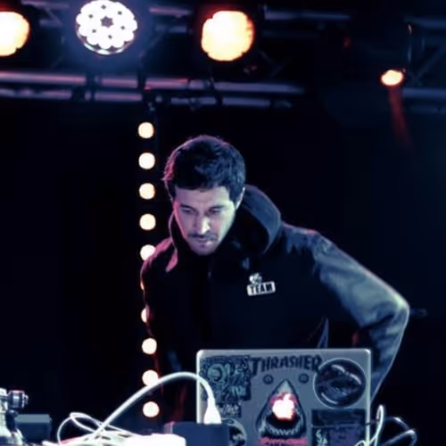 Kool Trasher performing at a DJ setup, intensely focused on his laptop covered with stickers, under vibrant stage lighting. He wears a dark jacket, blending into the moody, dimly lit background.
