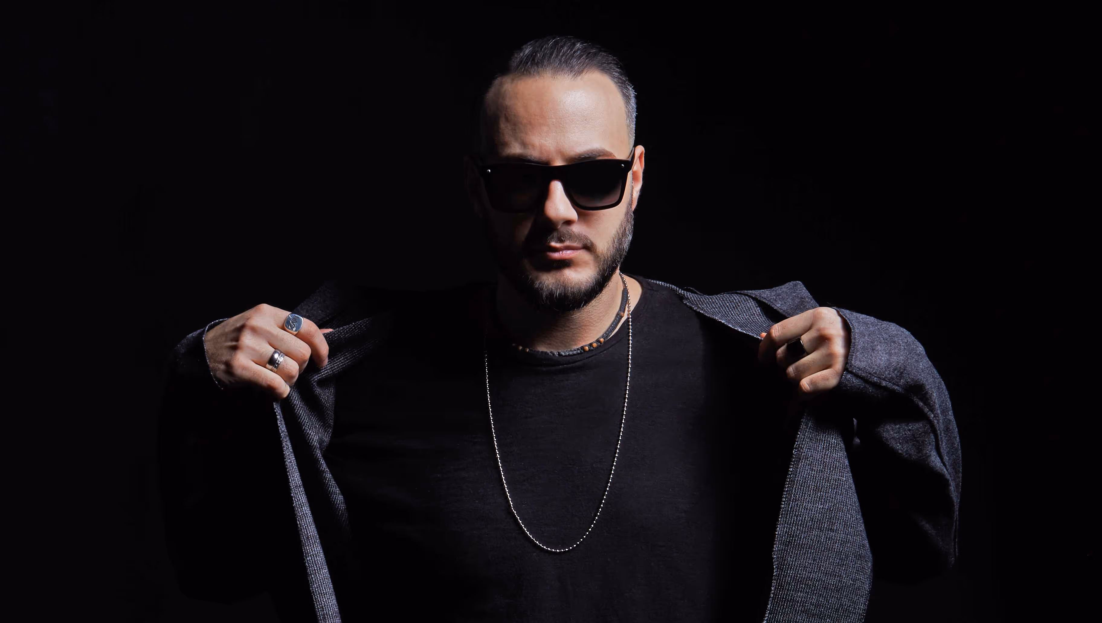 Sisko Electrofanatik posing against a dark background, wearing sunglasses and a chain necklace. He holds his jacket open with both hands, showcasing a confident and intense expression.