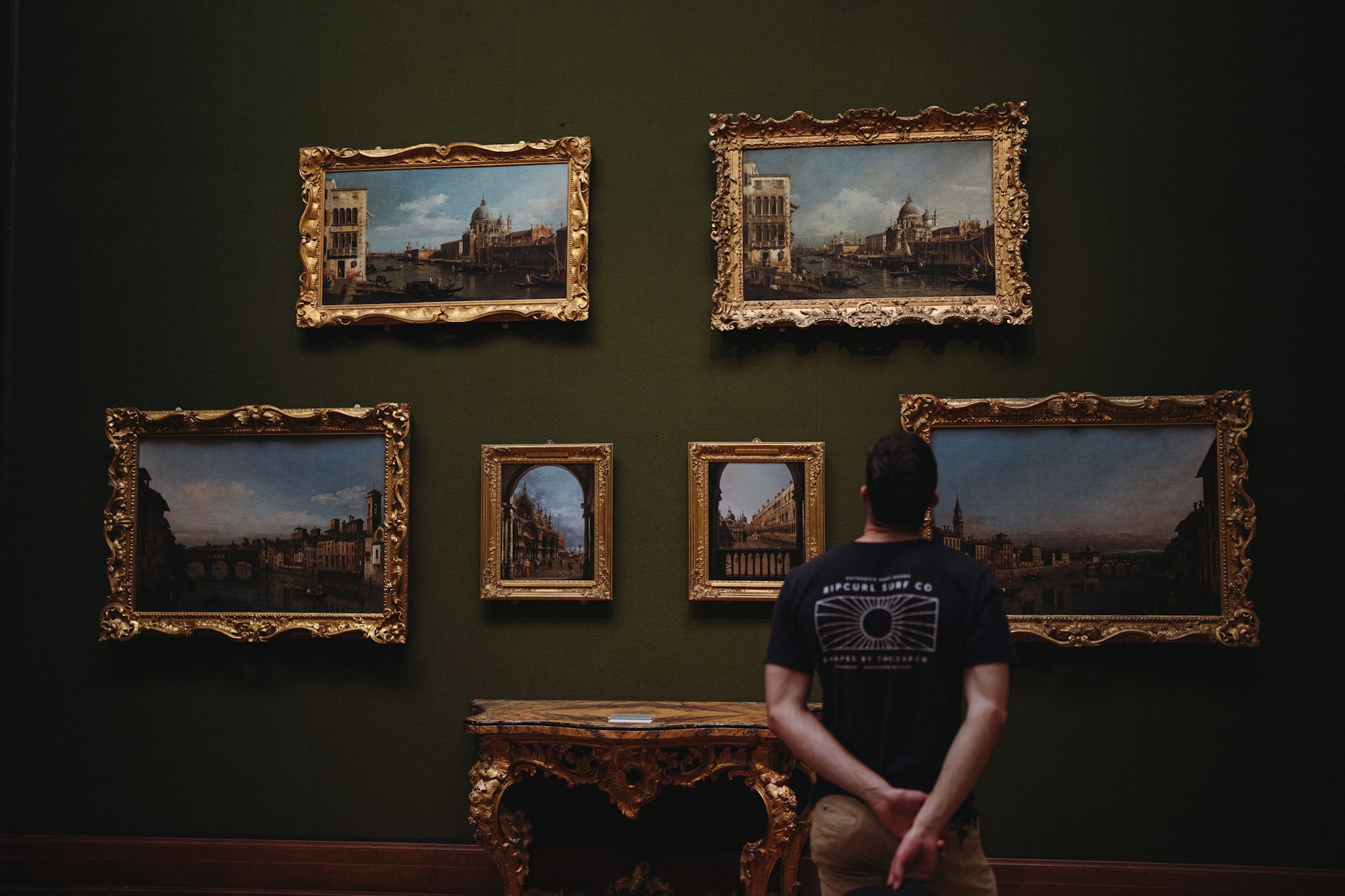 Visitor observing six framed paintings in a gallery with dark walls.