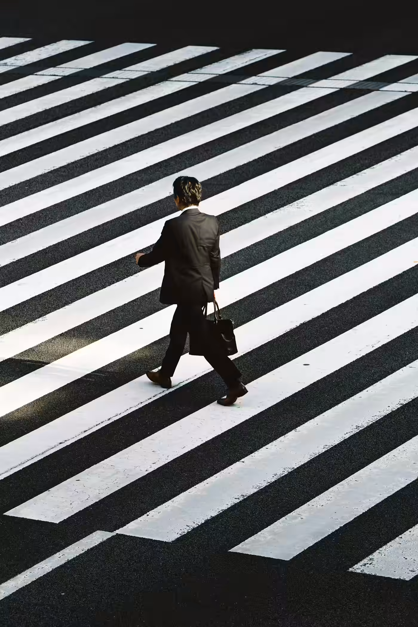 Homme en costume noir traversant un grand passage piéton à rayures blanches et noires.