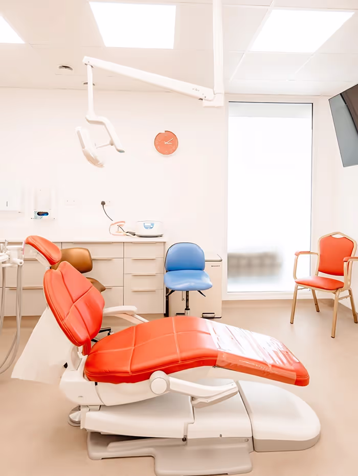 Modern dental clinic room featuring an orange dental chair, blue assistant stool, and orange visitor chair under bright lighting.