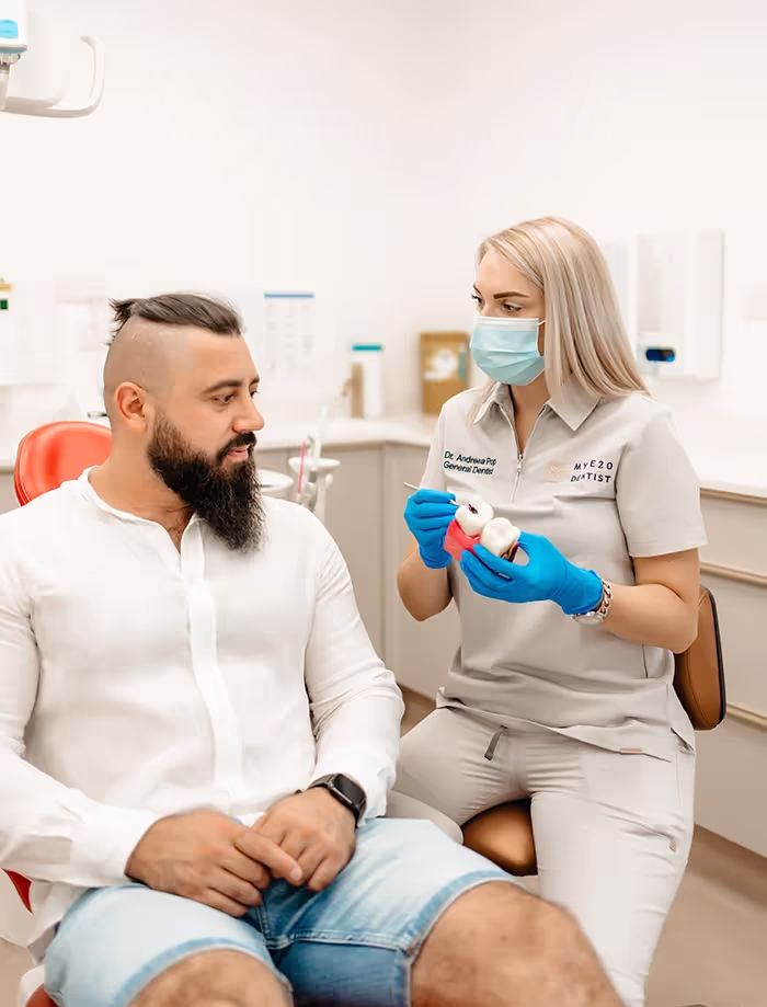 Female dentist wearing blue gloves and face mask explaining a dental model to a seated male patient in a dental office.
