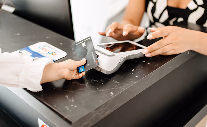 Person holding an American Express credit card near a contactless payment terminal on a black marble counter.