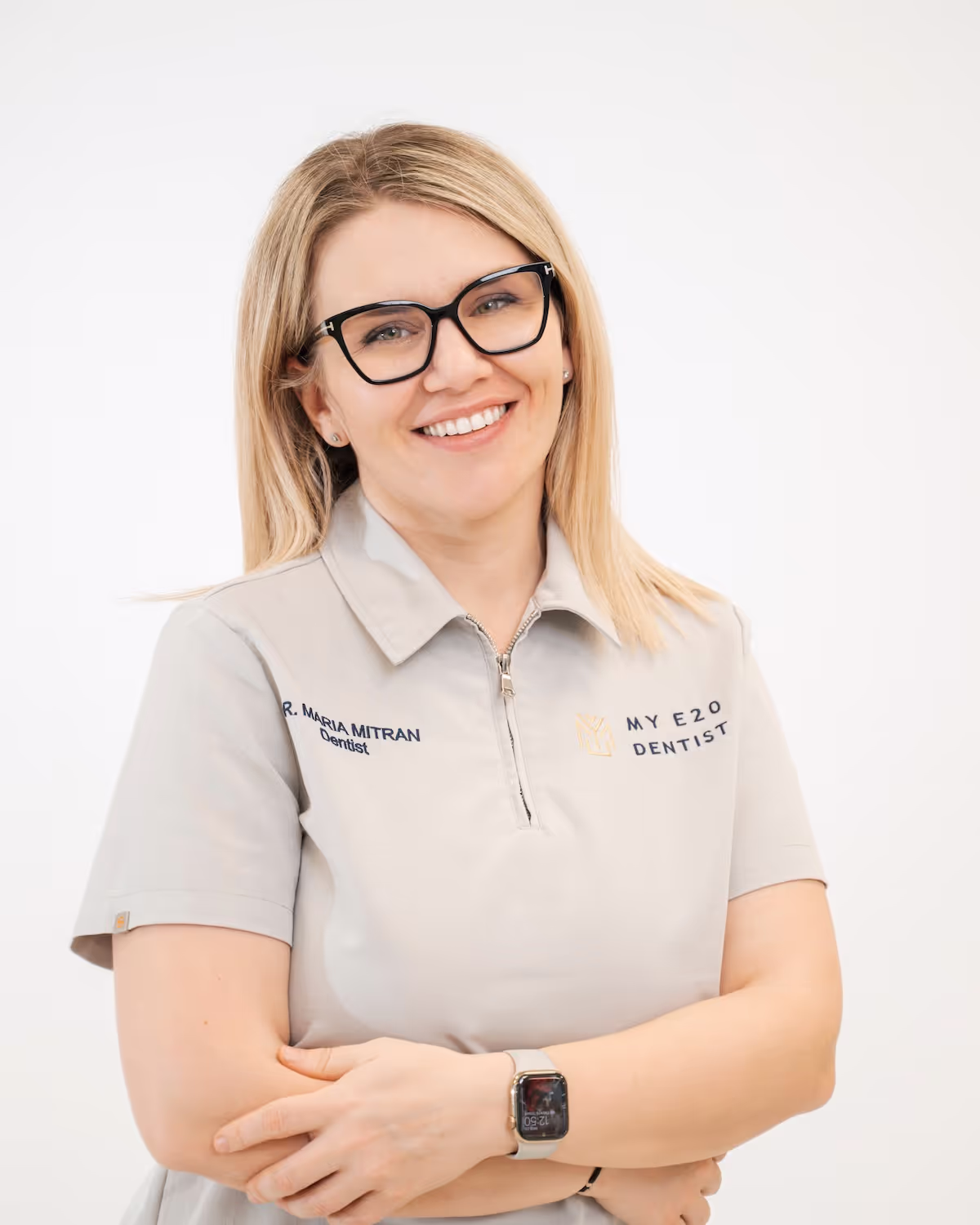 Smiling female dentist with blonde hair, black glasses, and a gray polo shirt embroidered with 'Dr. Maria Mitran Dentist' and 'My E20 Dentist.'