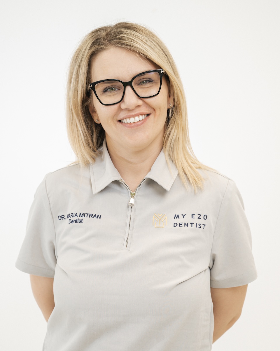 Smiling female dentist with blonde hair, wearing glasses and a gray uniform embroidered with the name Dr. Maria Mitran and MY E20 Dentist.