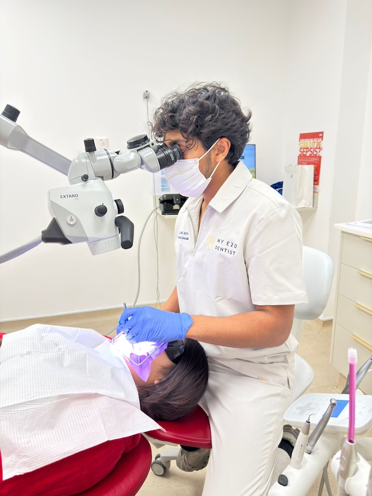Dentist wearing a mask and gloves using a microscope to examine a patient lying in a dental chair.