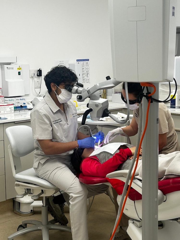 Dentist wearing mask and gloves using microscope to treat patient reclining in dental chair.