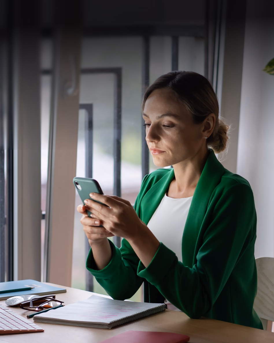 Professional woman in green blazer reviewing smartphone at desk with notebook and papers