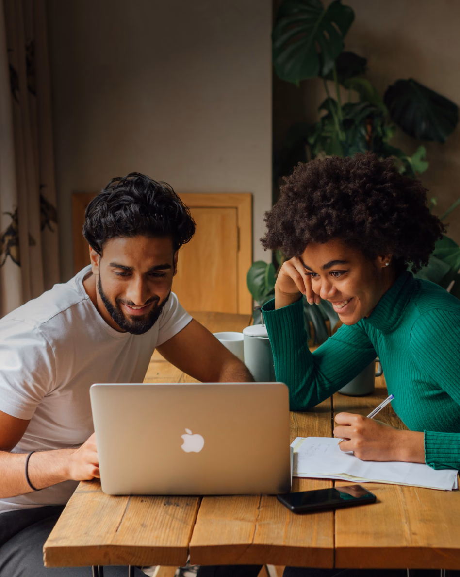 Two people collaborate at a wooden table with a laptop, notebook, and coffee cup in a bright room with plants