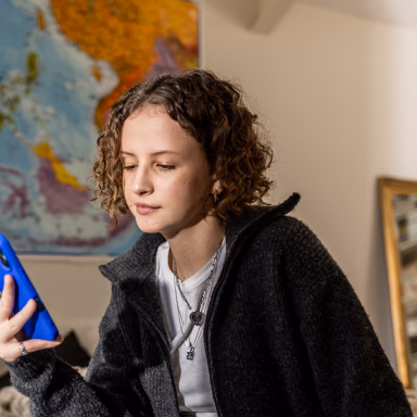 Woman with curly brown hair wearing a black cardigan and cross necklace, holding a blue object in an indoor setting