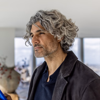 Man with gray curly hair wearing a black jacket over navy shirt in a modern interior