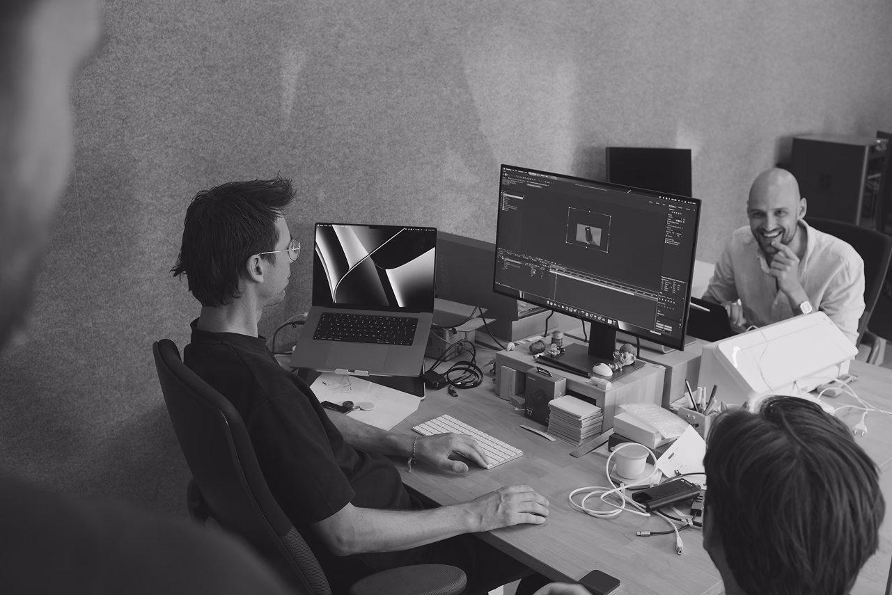 Three people collaborate at a desk with dual monitors in a black and white office setting