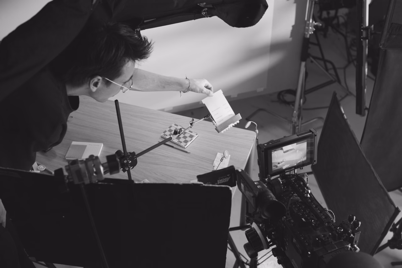 Person wearing glasses working at a desk with camera equipment and papers in black and white
